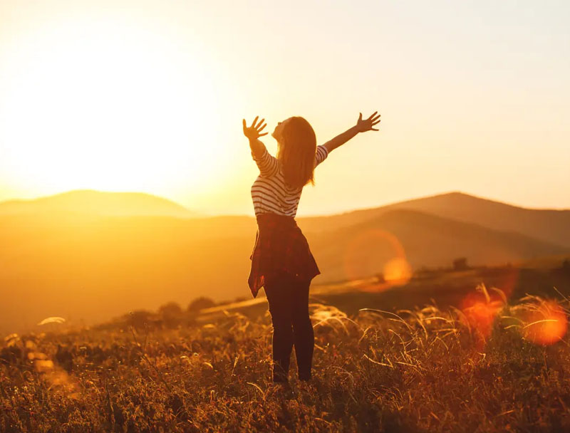 Selbstbewusst Frau streckt die Arme in die Höhe, hinter ihr der Sonnenuntergang